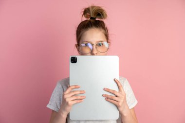 Çekici, tabletli ve pembe arka planda gözlüklü sarışın kız. Sevimli bir genç tablet kullanıyor, yüzünü tabletle kaplıyor, sadece gözleri görünüyor..
