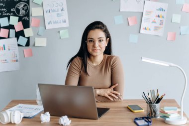 Masada dizüstü bilgisayarla çalışan gözlüklü güzel bir kız. Gri arkaplanda bir sürü renkli etiket var..