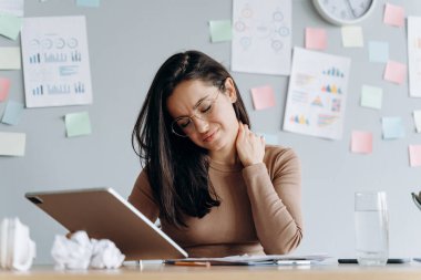 Tablet üzerinde çalışmaktan yorgun düşmüş, boynuna dokunmuş, yorgunluktan gözlerini kapatmış. Masadaki genç ve güzel kadın modern bir ofiste çalışıyor..