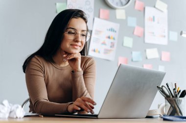 Genç memnun, mutlu, güzel iş kadınının görüntüsü ofiste dizüstü bilgisayarla oturuyor. Kız gözlük takıyor..