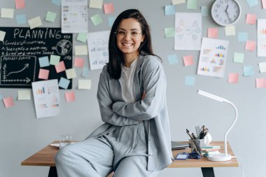 Masaüstüne oturmuş gözlüklü güzel bir esmer. Ofisin arka planında gülümseyen kız..