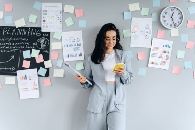 Üzerinde çıkartmalar olan gri bir duvarın arka planında elinde telefon tutan kendine güvenen bir esmer. Ofiste çalışan özenli, genç bir kadın..