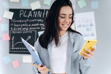 Ofisteki gri bir duvarın arka planında güzel bir esmer. Elinde önemli belgeler ve bir telefon var. Gri duvarda iş planlaması için bir tahta var..