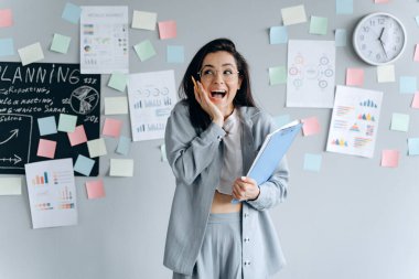 Elinde belgelerle duvarın arka planında raflarla yapıştırılmış harika bir iş kadını. Planlama tahtası. Ofisteki sevimli, güzel kız..