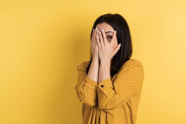 Çekici kız elleriyle yüzünü kapatıyor, sadece gözleri görünüyor. Turuncu arkaplanda izole edilmiş, metin için yer.