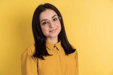 Sevimli, gözlüklü, turuncu duvarın arkasına poz veren esmer bir kız. Boş bir duvarda izole edilmiş, yakın çekim.