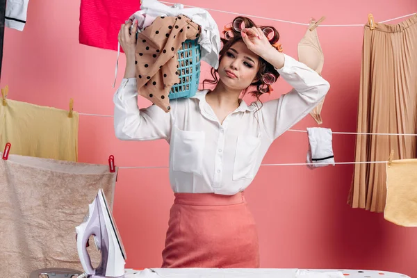 Ev hanımı kirli çamaşırlarla dolu bir leğen tutuyor, ıslak giysileri çamaşır ipine asıyor. Yüzü hoşnutsuz, pembe arka planda soyutlanmış. Oda servisi kavramı