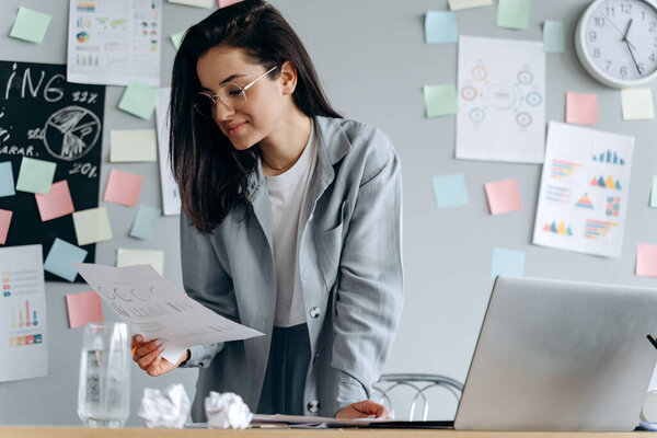 Милая, деловая девушка в очках работает в офисе, она тщательно изучает документы. На заднем плане офиса милая молодая женщина, работающая.