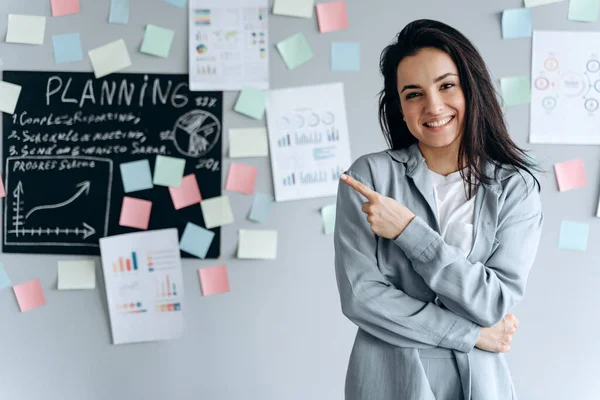 Üzerinde çıkartmalar olan gri bir duvarın arka planında gülümseyen genç bir kız. İşaret parmağıyla bir şeye işaret ediyor..