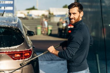 Yüksek basınçlı köpüklü çamaşır makinesiyle arabasını yıkayan adamın portre görüntüsünü büyüt. Kameraya gülümseyen zevk erkeği 