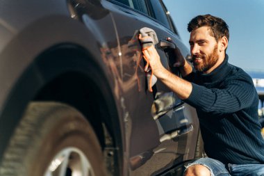 Adam sokakta arabasını siliyor. Sıradan giyinen yakışıklı sakallı adamın yakın görüntüsü araba kapılarını ve kaputu mikrofiber bezle yıka. Güneşli bir günde araba yıkama konsepti 