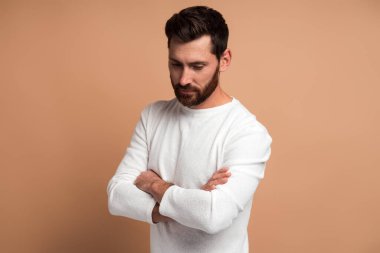 Çapraz kollu yakışıklı, dalgın sakallı genç bir adamın bej arka planda kameranın önünde poz verirken düşünceli bir ifadeyle aşağı bakan portresi. 