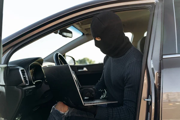 Masked thief in a balaclava stealing laptop from car and looking at the screen while sitting inside. Criminal concept. Stock photo