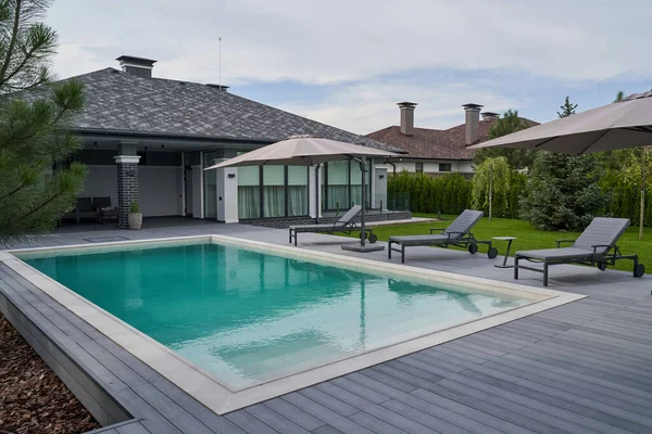 Wooden lounge chairs in modern villa pool and deck. Light blue water swimming pool of a luxurious mansion with chairs around it. Modern house at the background