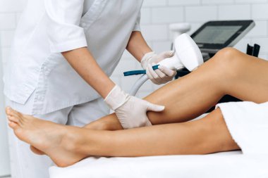 Estetik vücut tedavisi. Kırpılmış kafa portresinin yanında beyaz bornozlu, rahat genç bir kadın diz bölgesindeki bacaklarının kıllarını aldırma prosedürü uyguluyor. 