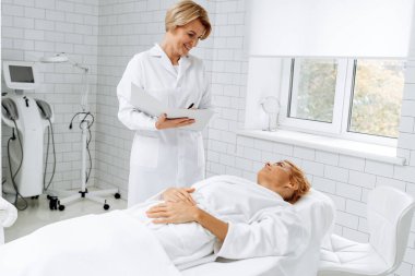 Beyaz bornozlu çekici bir kadının portresi. Yatakta uzanmış, gülümseyerek güzellik uzmanına bakıyor. Onunla konuşurken ve klasörü tutarken. 