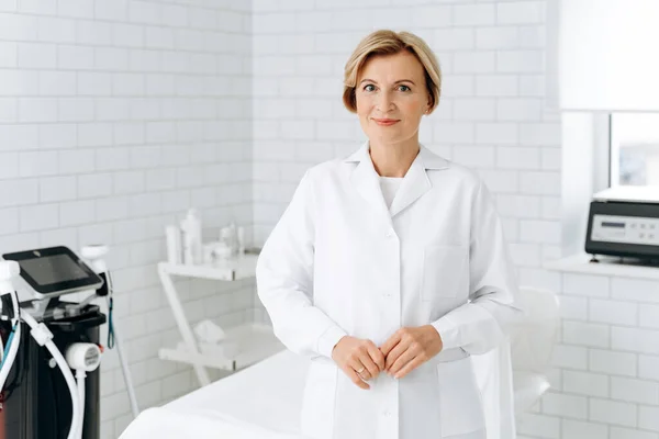 Çarşafla örtülü daybed yakınındaki ayakta beyaz önlük içinde büyüleyici güzellik portresi. O kameraya bakıyor ve gülümsüyor 