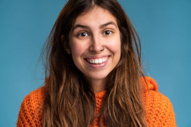 Mavi bir duvarda duran ve samimi bir şekilde gülümseyen güzel, mutlu bir genç kadının portresi. Modern kız poz verirken kameraya gülümsüyor.