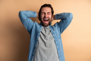 Portrait view of angry or shocked handsome bearded young man in blue casual style shirt standing and screaming. with closed eyes. Indoor studio shot, isolated on orange background 