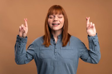 Fortune please come. Portrait of woman standing with crossed fingers for good luck, heartily making wish, praying and hoping for miracle. Indoor studio shot isolated on orange background 