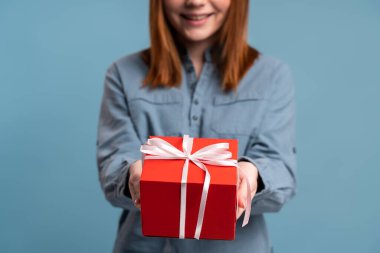 Smiling woman holding out present box, giving gift, looking at camera with optimistic expression, greeting. Indoor studio shot isolated on blue background 