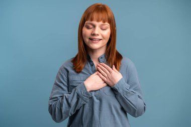 Head shot portrait happy ginger woman smiling and looking at camera while holding hands on chest and feeling gratitude. Gesture of sincere feelings from heart and love concept 