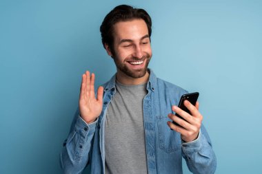 Portrait of stunning happy young adult man waving with hand, looking seductively while has online conversation on smartphone. Indoor studio shot isolated on blue background 
