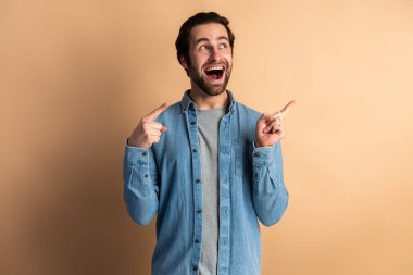 Portrait of handsome happy man in stylish denim shirt pointing aside, showing blank copy space for idea presentation, commercial text. Indoor studio shot isolated on orange background 
