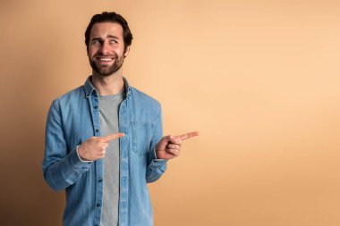 Cheerful hipster guy pointing at empty place for commercial idea, looking away with toothy smile. Studio shot isolated on orange background 
