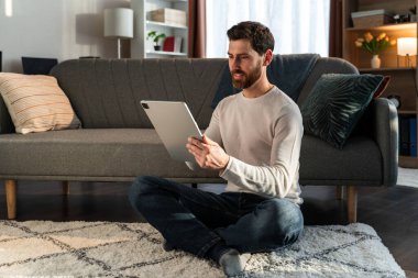 Cheerful young male is sitting at the living room at the lotus pose and looking at tablet screen in hands with interest 