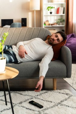 Bearded man sleeping on sofa with tv remote control at hands at home during the day. Leisure and people concept 