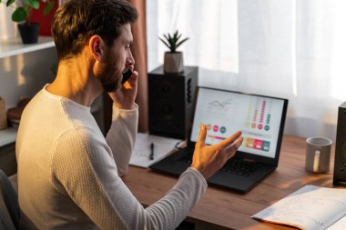 Back view of the serious male sitting in front of his laptop and looking at the screen while chatting via smartphone with clients 