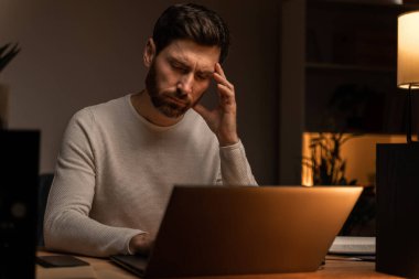 Young male is feeling exhausted after using laptop late at night in living room. Guy looking at the screen with tired face 