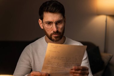 Bearded man wearing glasses reading interesting letter with serious expression while spending evening alone at home 