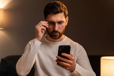 Handsome young male smiling while sitting with smartphone and looking attentively at the screen. Stock photo