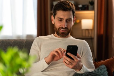 Online chat. Smiling young man at home using modern smartphone. Brunette guy checking messages while sitting at the sofa. Domestic life concept 