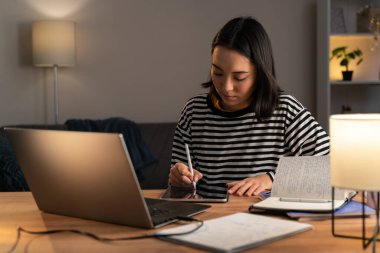 Asyalı kadın tasarımcı akşam evde dizüstü bilgisayarında çalışırken tablette bir şeyler çiziyor. Stok fotoğrafı
