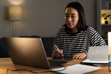 Laptop bilgisayarıyla masada otururken tablette resim çizen güzel bir genç kadın. Evde çalışırken ekrana bakan sevimli kız.