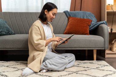 Genç bir kadının tablet ekranda oturup yazışının tam boy görüntüsü. Sosyal ağda arkadaşlarıyla sohbet ederken tableti elinde tutuyor.