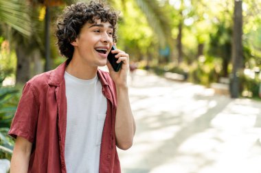 Kıvırcık neşeli adam şehir caddesinde yürüyor ve akıllı telefon kullanan biriyle sohbet ediyor. Aktif ve mutlu. 