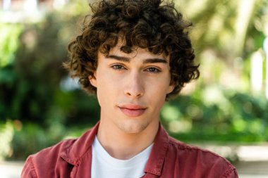 Yeşil yaz parkında yürürken sakince kameraya bakan mutlu kıvırcık adamın portresi. 