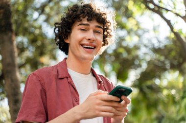 Kıvırcık adam güneşli sokakta dinleniyor ve akıllı telefonunu elinde tutarken mutlu bir gülümsemeyle kameraya bakıyor. 