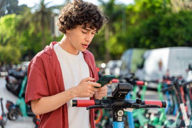 Konsantre genç adam yaz günü ulaşımını açarken şehrin yakınındaki kick scooter istasyonunda uygulama kullanıyor. 