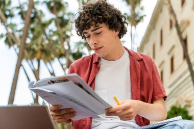 Kentteki bankta öğrenirken notlara dikkatlice bakan modaya uygun giyinmiş erkek öğrencinin portresi 