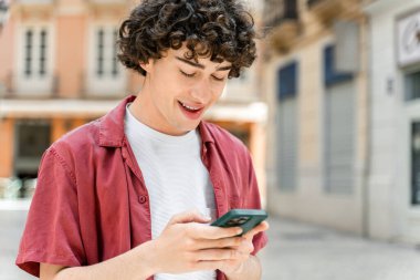 Kıvırcık, mutlu, sıradan kıyafetler giyen genç bir adam sokakta dikilip akıllı telefonuna zevkle bakıyor. 