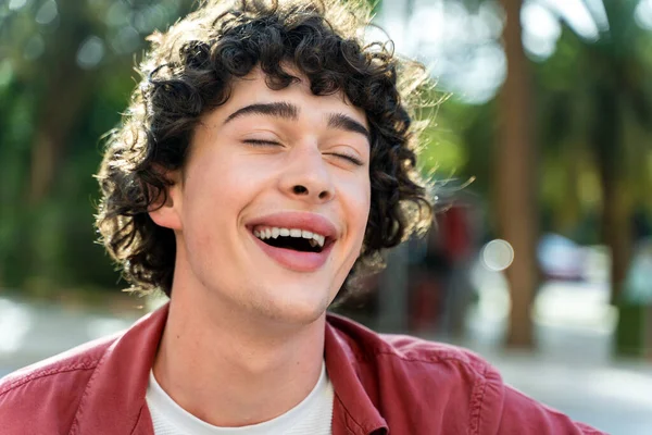 Portrait of a happy curly man laughing out loud and relaxing with closed eyes while sitting on the bench at the park. Stock photo