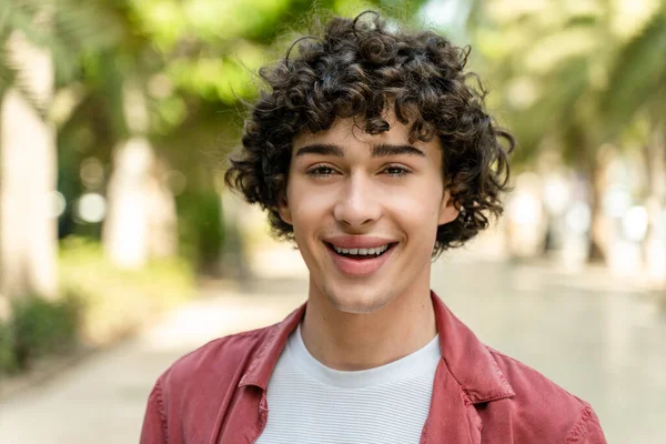 Portrait of cheerful young curly man standing in city park on sunny warm day and smiling at the camera. Stock photo