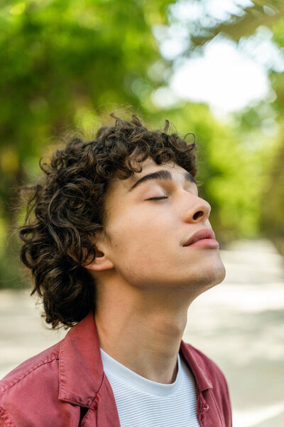 Вертикальный вид красивого расслабляющего молодого человека, стоящего с закрытыми глазами и улыбкой удовольствия во время прогулки на свежем воздухе 