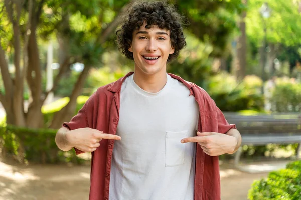 Young curly man standing at the street looking confident with smile on face, pointing oneself with fingers proud and happy