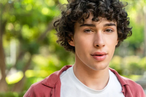 Portrait of cheerful young curly man standing in city park on sunny warm day and smiling at the camera. Stock photo